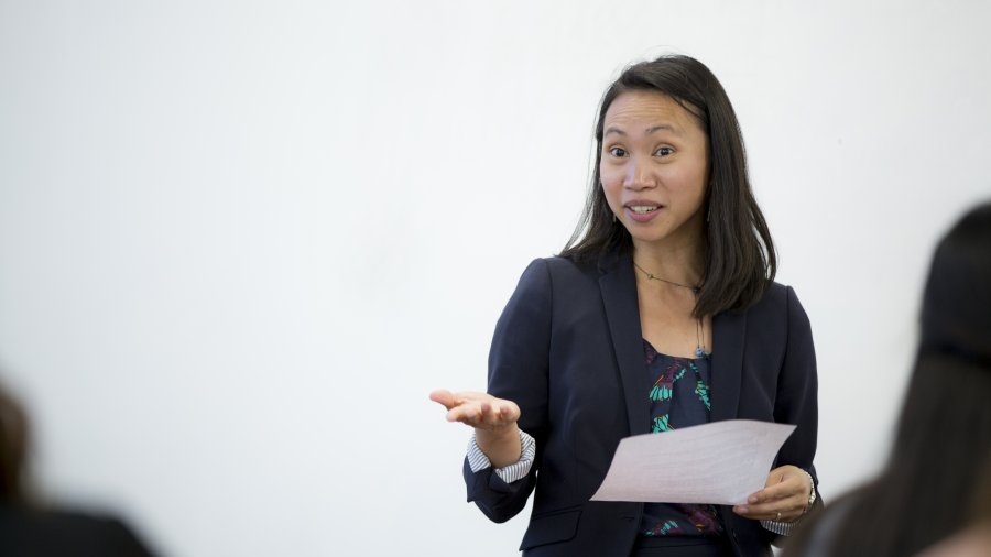 A woman in a black blazer is holding a piece of paper and speaking as she stands in front of a white board.