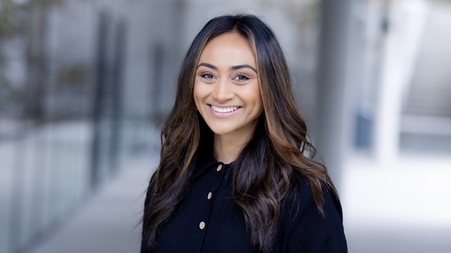 A smiling woman with long, wavy dark hair wearing a black buttoned top poses for a professional portrait.