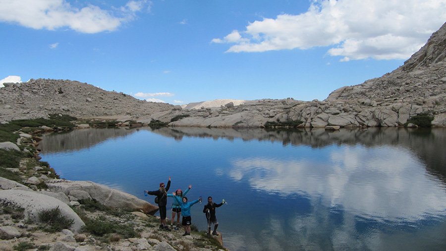 A group of students raise their hands in celebration on a hike near a clear lake.