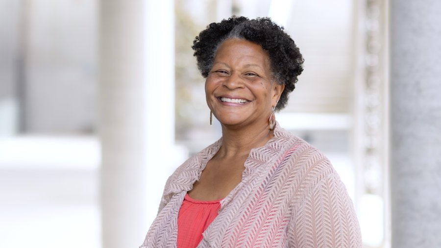 A smiling woman with short coily hair wearing a coral top and light patterned cardigan poses for a professional portrait.