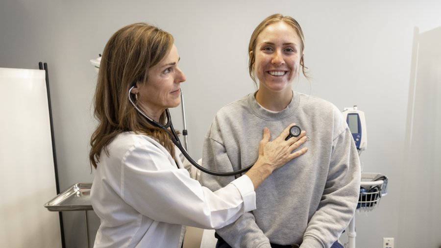 A female nurse with brown hair is holding a stethoscope to her patient's heart. The patient is a female with blonde hair. She is sitting on the exam table, smiling.