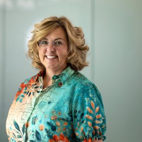 A smiling woman with shoulder-length curly blonde hair and glasses wearing a colorful floral blouse poses for a professional portrait.