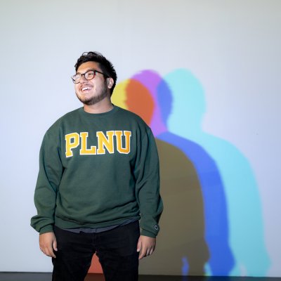 Alum Josue Barragan poses for a photo smiling in front of white background and graduate's shadow formed in colorful hues.