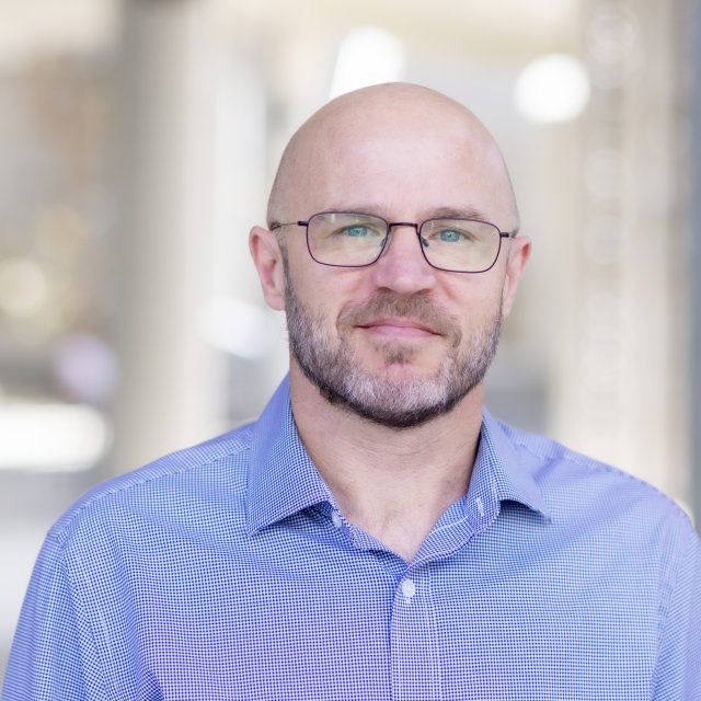 A man with a shaved head, short beard, and glasses wearing a blue collared shirt poses for a professional portrait.