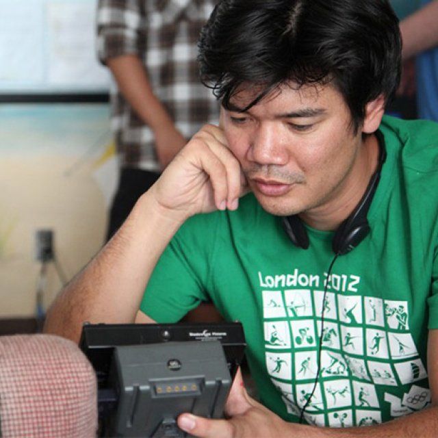 Male in green shirt and headphones around his neck looks into a camera viewfinder.
