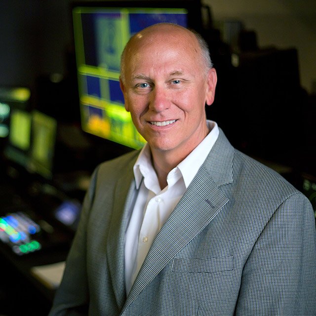 Alan Hueth stands in front of production studio screens and communication device technology.