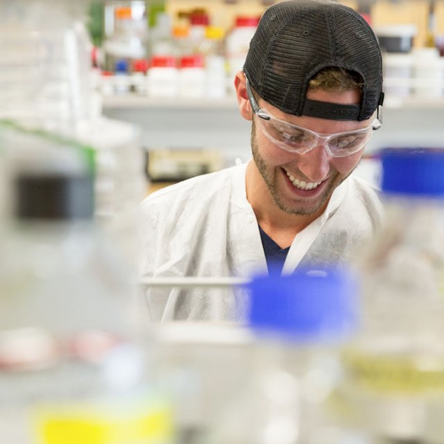 A view of a male student smiles as he looks down at his lab work through the bottles on the lab shelves