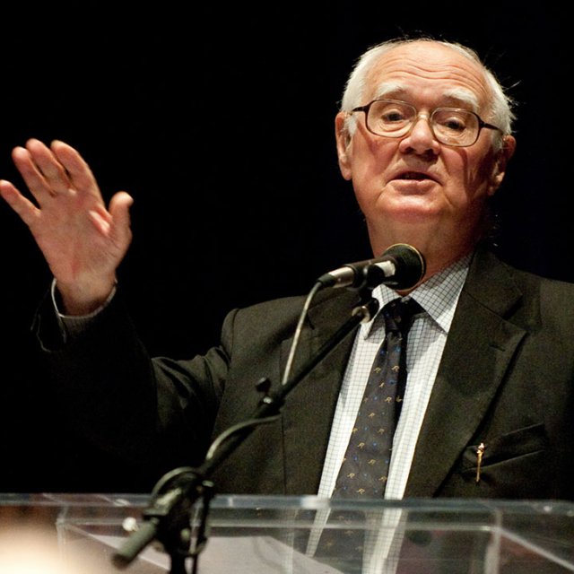 Polkinghorne, an older man, speaks from a podium to community in Crill Hall at PLNU