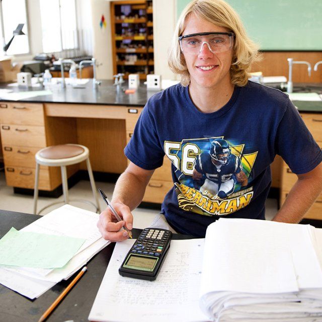 A male student wearing lab safety glasses looks up and smiles from calculating a physics equation.