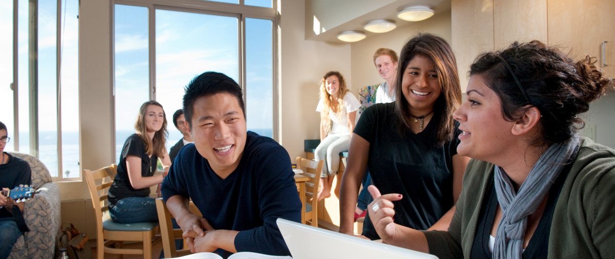 students gatherd around talking. one student has her laptop open. another student has his textbook open.