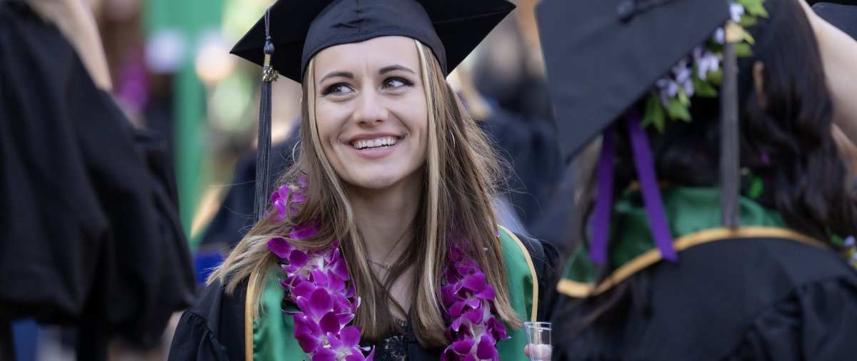 A female student with brown hair is smiling at her PLNU graduation ceremony. She is wearing a black cap and gown, as well as a purple lei.