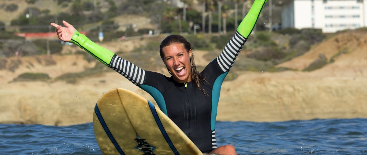 Young woman wearing a wetsuit sits on a surfboard in the ocean with her hands above her head in excitement