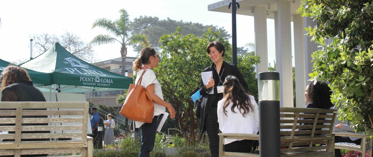 Attendees talk near research displays and PLNU tents during an outdoor academic event on campus.