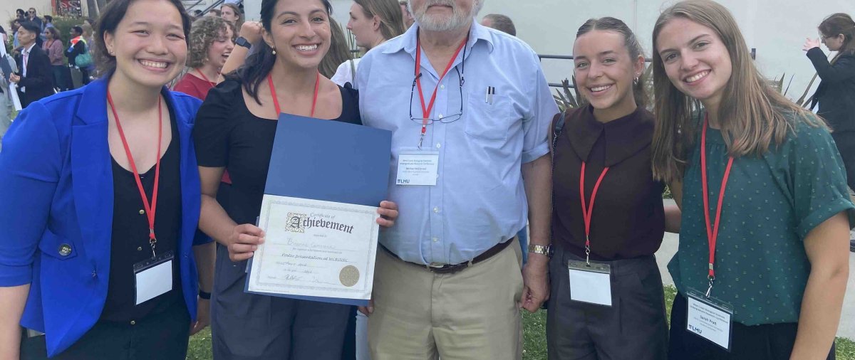 Four students and a faculty member pose together at an academic conference, with one student holding an achievement certificate.