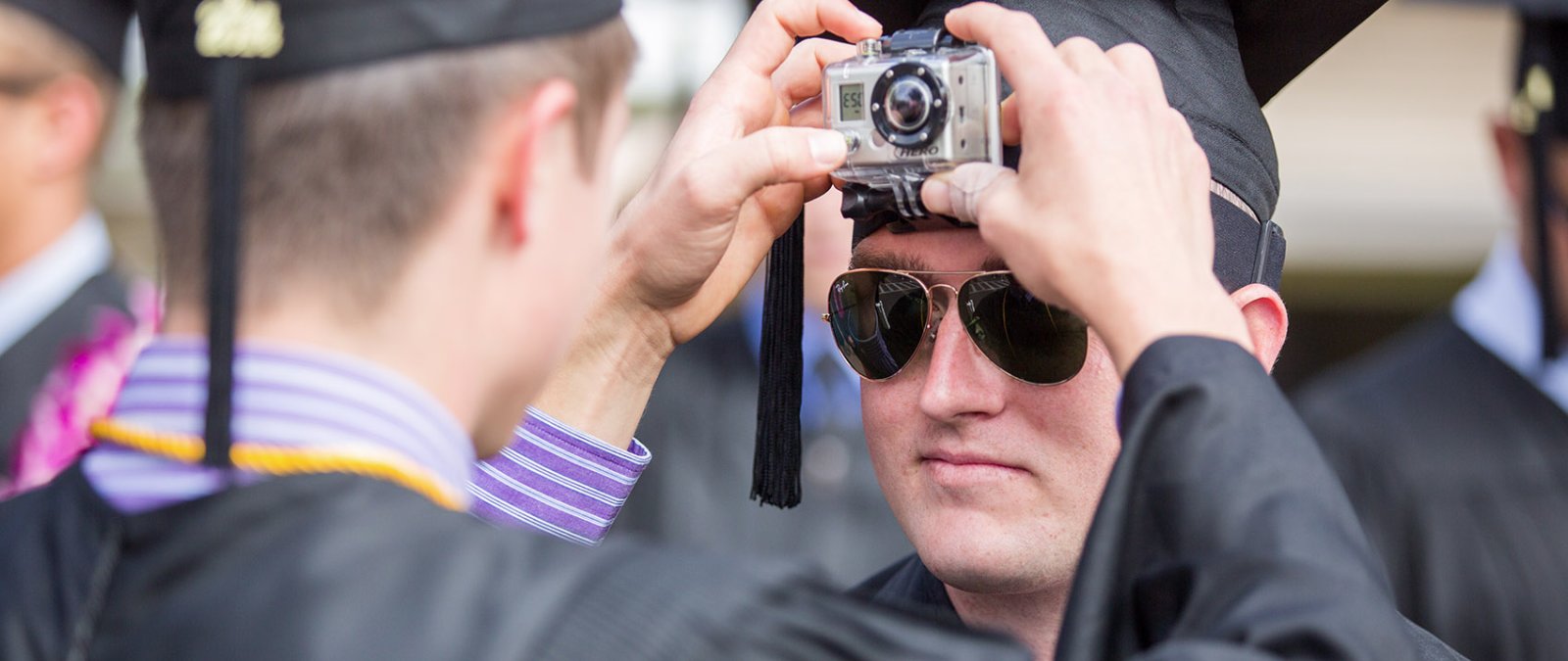 A PLNU student helps his friend turn on a GoPro attached to his graduation cap.