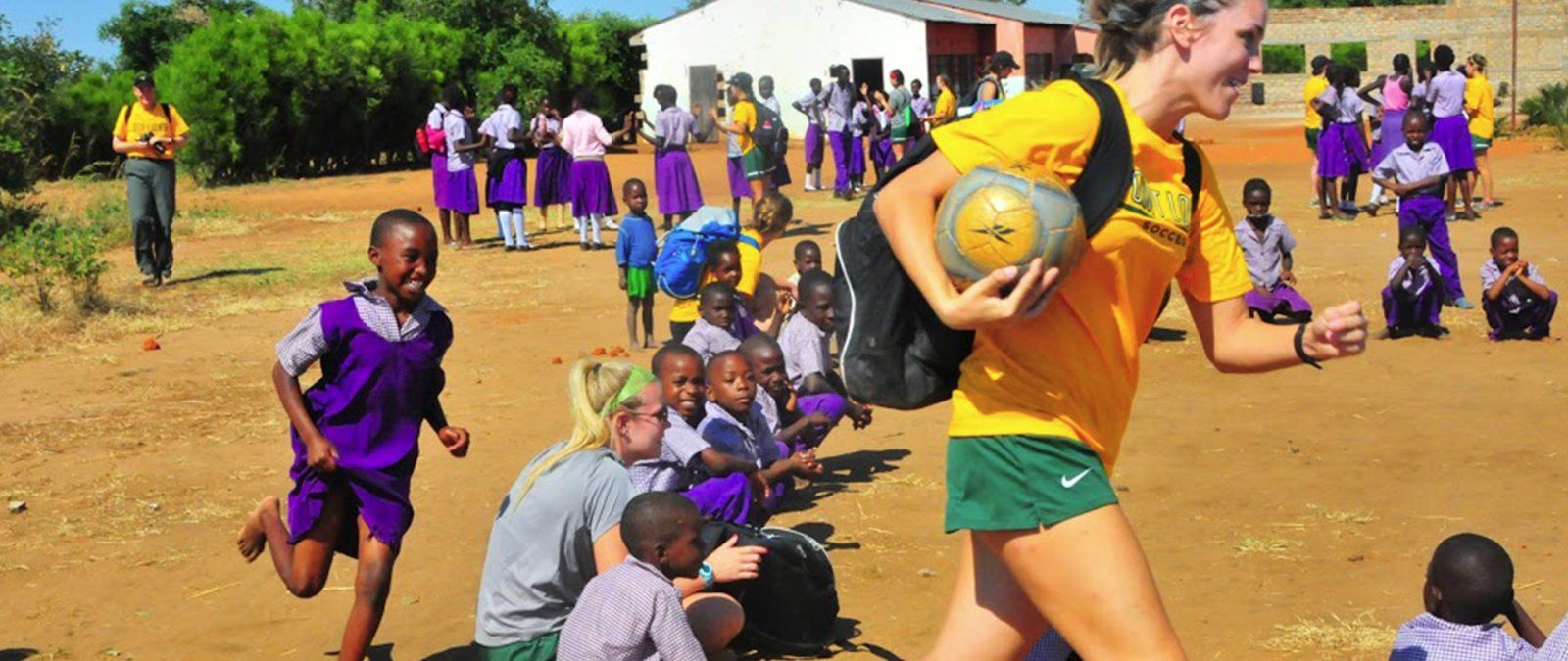 A female soccer player carrying a soccer ball is chased by a child in Zambia around a circle.
