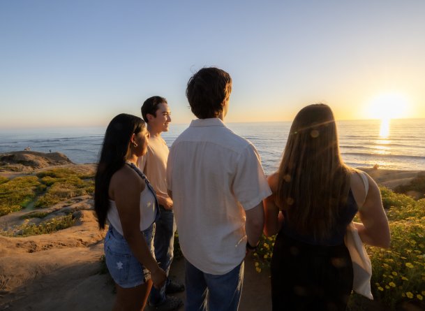 Four students stand on Sunset Cliffs together. They are smiling and looking at the sunset in the horizon.