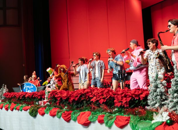 several college students singing into microphones, wearing christmas outfits and beach outfits. The backdrop is red and the stage is lined with red poinsettia flowers.