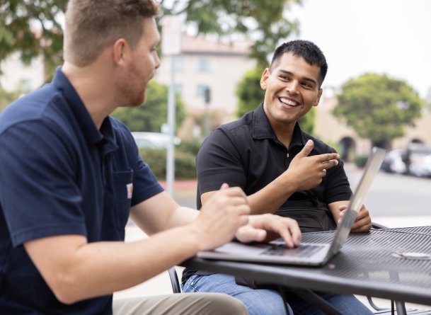 Two PLNU students work on laptops outside 