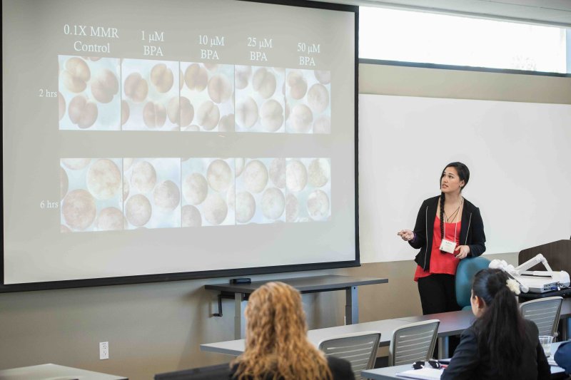 A presenter discusses scientific research displayed on a projection screen before an audience in a PLNU classroom.