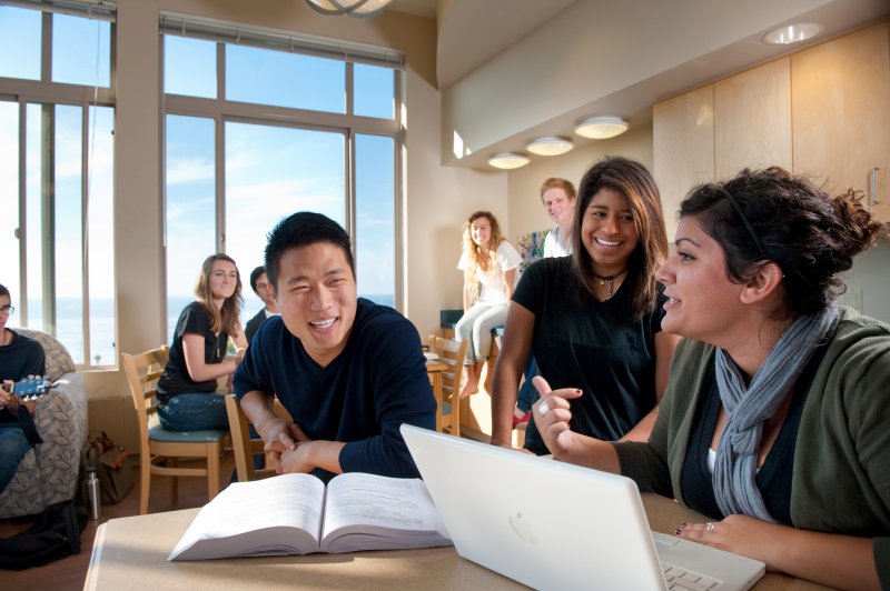 students gatherd around talking. one student has her laptop open. another student has his textbook open.