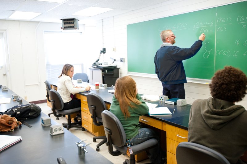 A male math teacher is writing on a chalkboard in front of his class. Behind him, his students are sitting at their desks, taking notes. 