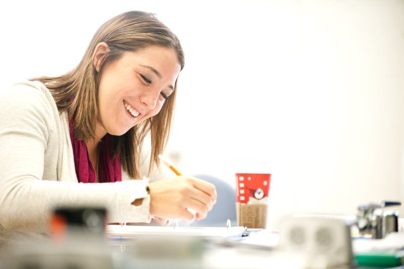 A female student smiles as she looks down at a paper on a desk. She is holding a pencil and is about to write.
