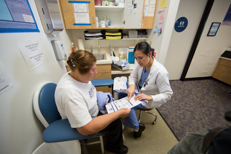 A female nurse in blue scrubs and a white coat is sitting across from a female patient, showing her a pamphlet. They are sitting in an exam room in a doctors office.