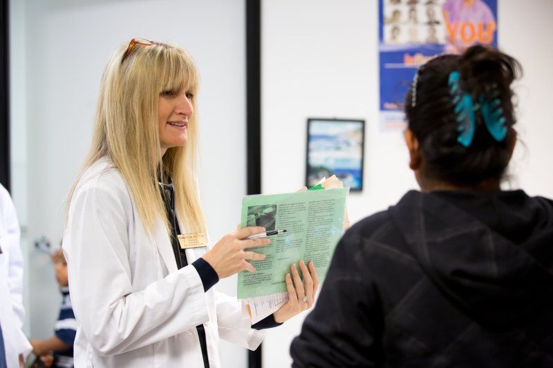 A female nurse with blonde hair is wearing a white coat over black scrubs. She is looking to the right, smiling, she talks to a patient. The patient, on the right, has black hair, a black hoodie, and blue hair clip.