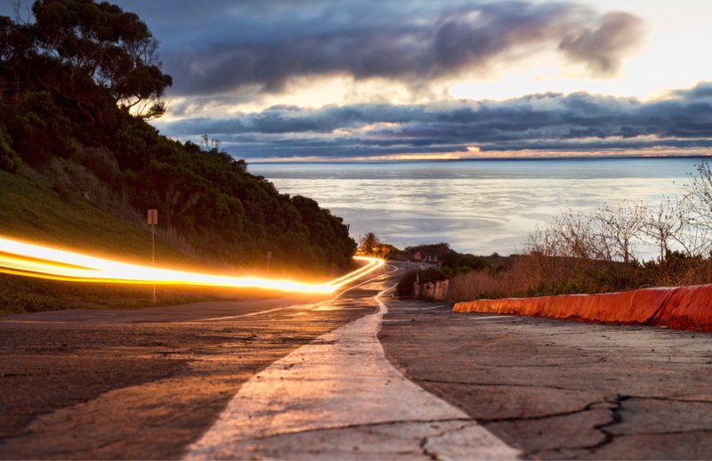 A street-level photo of the winding road down to Young Hall at night. The Pacific Ocean to the right and long exposure car lights to the left make up the composition.