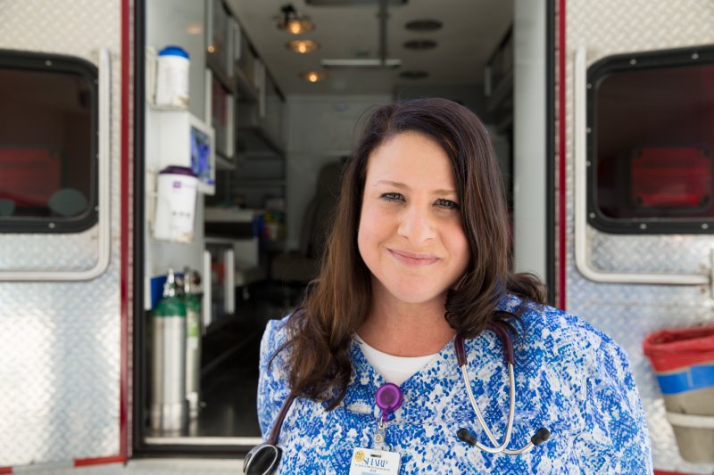A female nurse with brown hair is wearing blue patterned scrubs and has a stethoscope around her neck. She is smiling and is standing in front of the back of an ambulance.