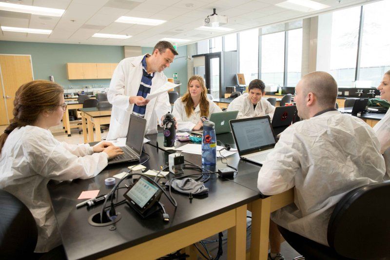 science professor and students all wearing white lab coats discussing a topic in a laboratory 