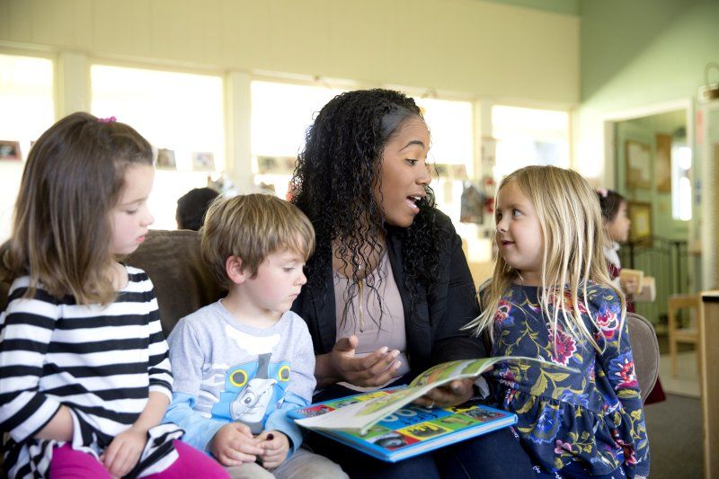 A female teacher is sitting, surrounded by three of her young students. She is reading them a book that is open on her lap.