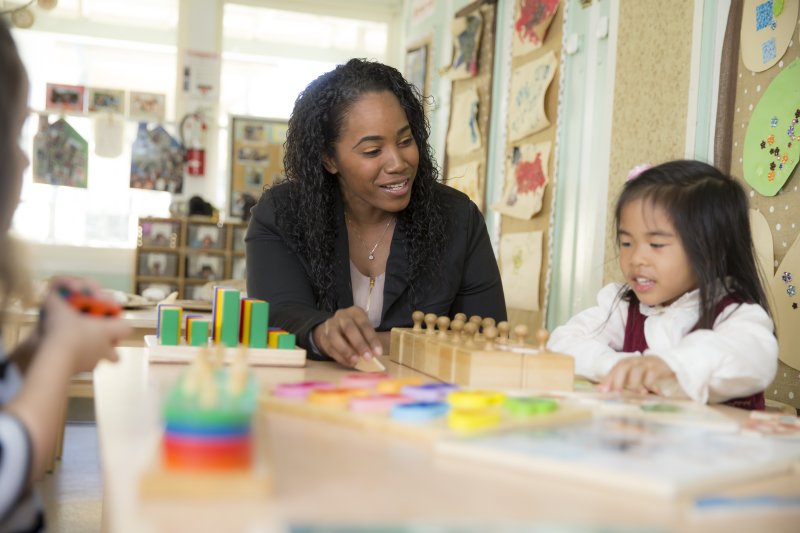 A teacher sits at a table with one of her young students. They are both smiling and are working on a puzzle together.