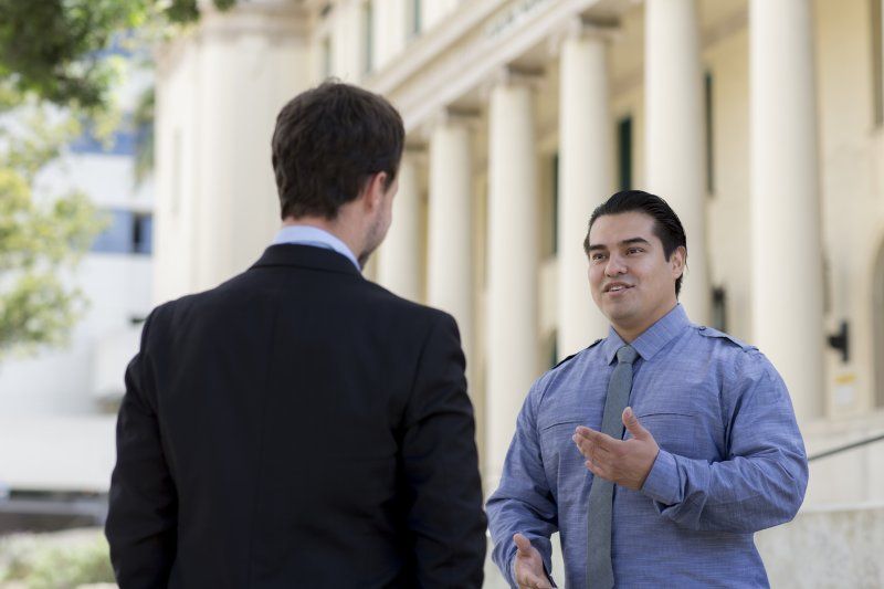 A man who works in public administration is standing outside of San Diego City Hall. He is wearing a blue button down and is talking to a man wearing a black suit.
