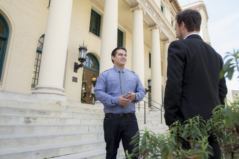 A man in a blue button down is standing in front of San Diego City Hall, talking to a man in a black suit.