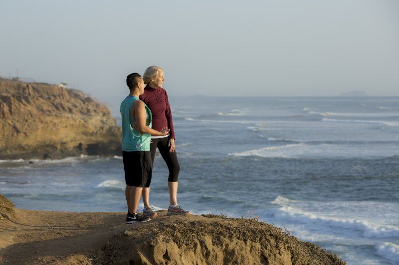 A man in a blue workout top and a woman in a maroon pullover stand next to each other on the side of Sunset Cliffs. They are talking while looking out at the ocean.