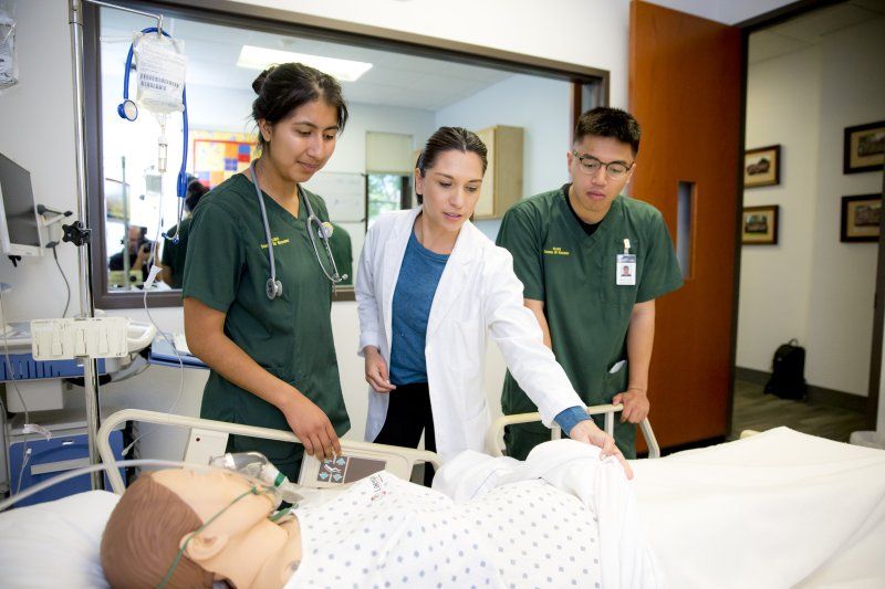 Two student nurses in green scrubs stand on each side of a nurse in a white coat. The three nurses are looking at a manikin laying on a hospital bed.