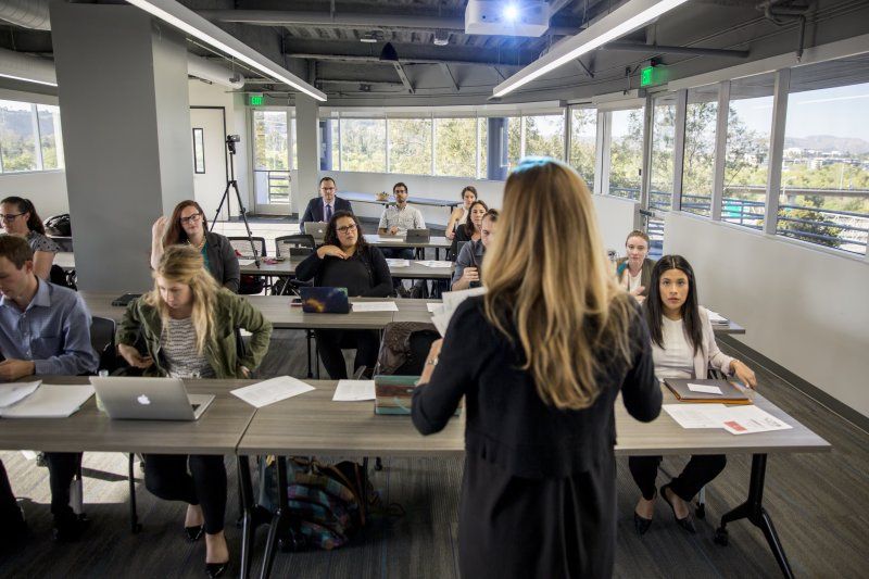 A writing professor with blonde hair stands in front of her class, giving a lecture. Her students are sitting, their laptops are open and writing papers on their desks.