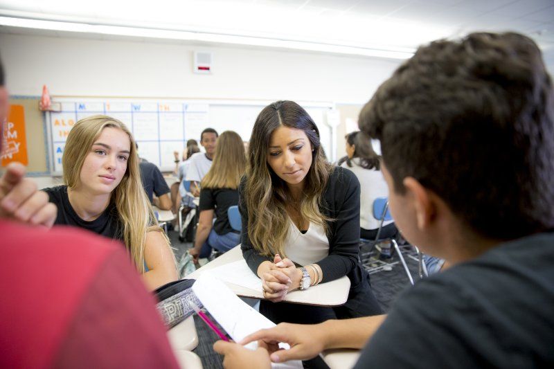 A female teacher in a black blazer and white t-shirt sits at a desk with three other students. She is looking at paper that a male student in a black shirt is holding.