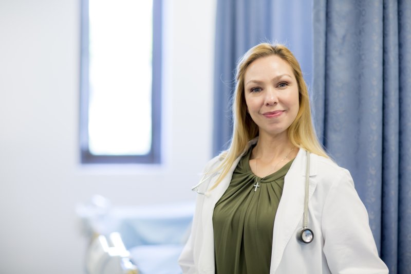 A female nurse with blonde hair is smiling. She is wearing a white coat and stethoscope around her neck.