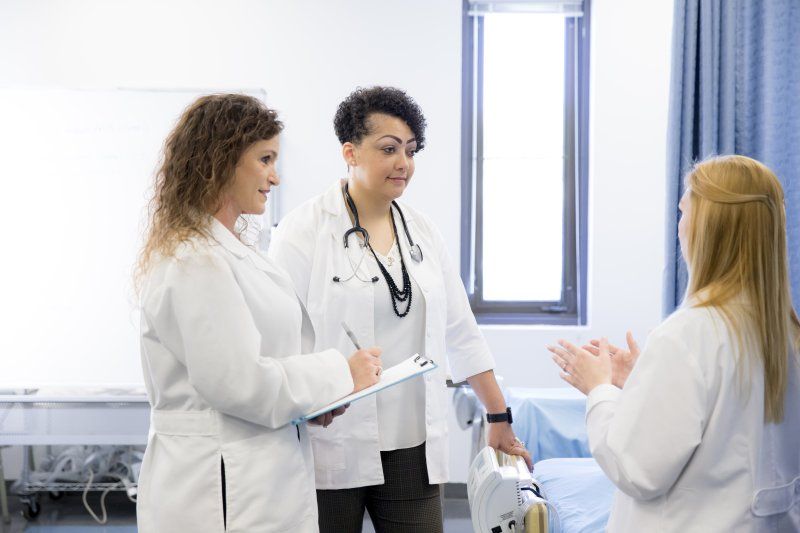 Three female Doctor of Nursing Practice (DNP) program members are discussing a patient's plans. They are all wearing white coats and are in a clinic.