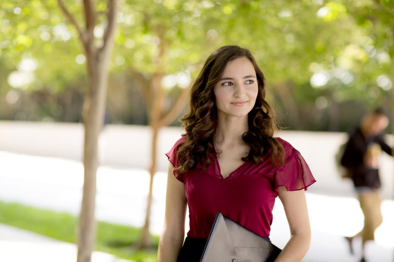 A woman in a red blouse is smiling to the right, holding a black folder. She is outside in a business center's courtyard.