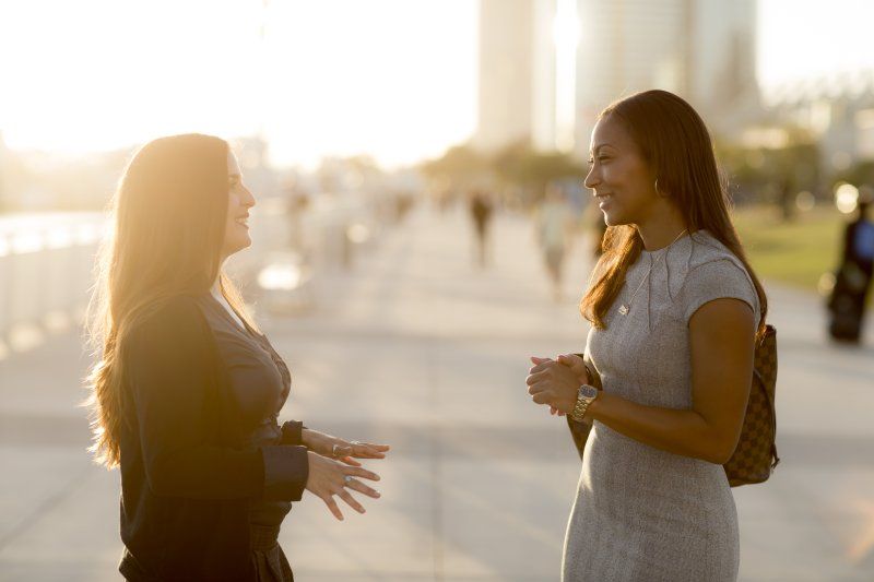 Two women stand outside, looking at each other while in conversation. The one on the left is in a black cardigan and the one on the left is in a grey dress.