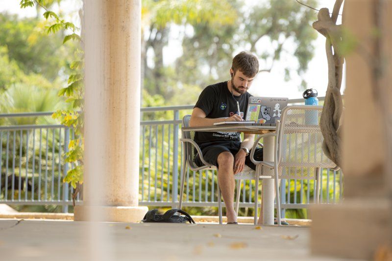 A male student is sitting at a chair and table at a covered balcony. His blue water bottle, an open laptop, and notebook are on the table. He is leaning forward and writing in the notebook.