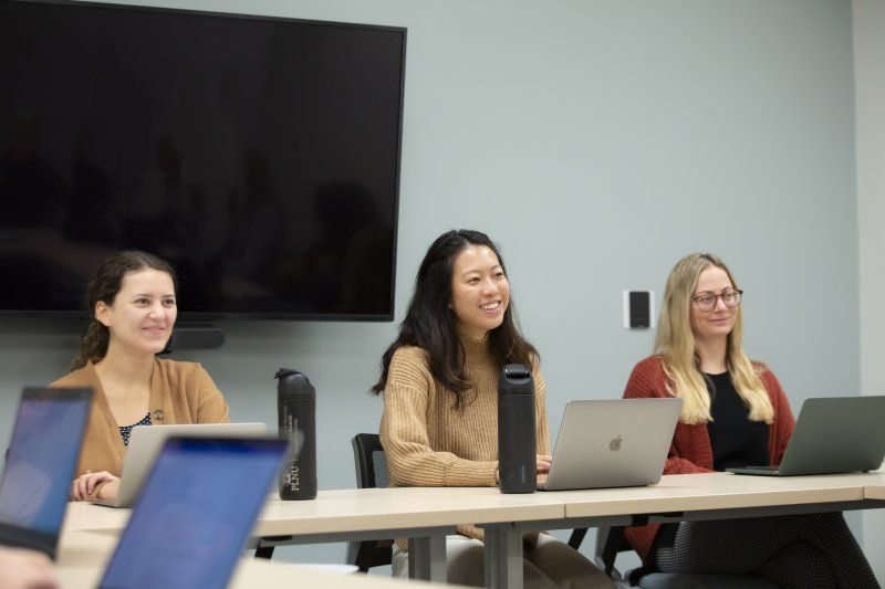Three female adult students are sitting at a desk, smiling. They are in a classroom and have their laptops open on the desk.