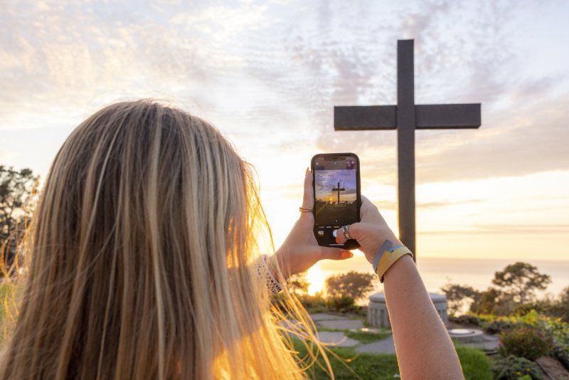 A girl is holding her cell phone up in front of her, taking a photo of a cross.