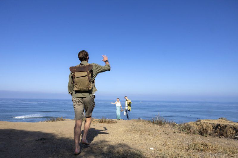 A male student with a green backpack is waving and walking toward two other students in the distance. They are standing outside with the ocean behind them.