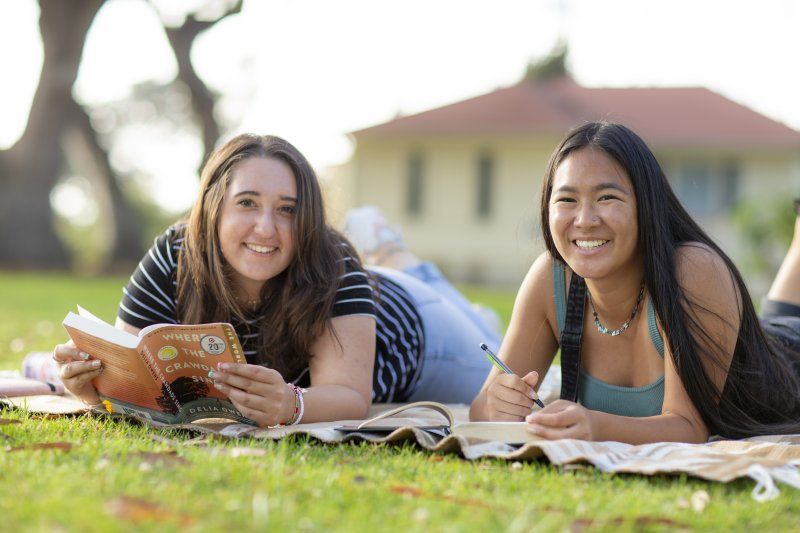 Two female students are laying on some blankets on a grass field. They are smiling, looking up from the textbooks that they're reading.