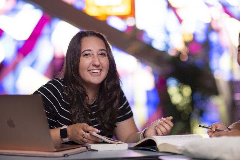 A female student is smiling. At the table in front of her, there are open books and is an open laptop.